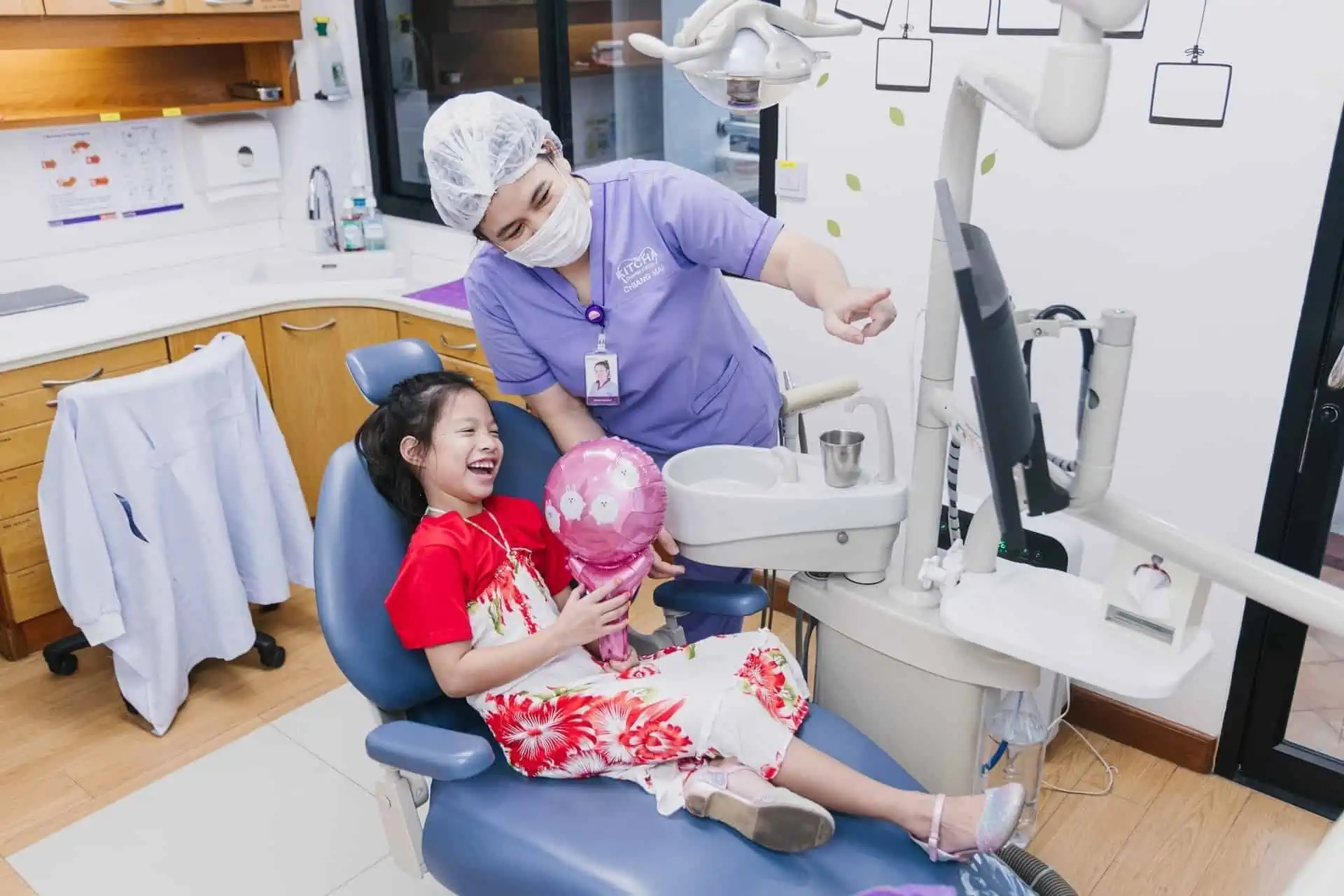 A young girl laughing happily in a dental chair at Kitcha Dental Clinic Chiang Mai with a cheerful atmosphere, receiving dental care from a professional dentist.